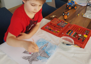 Chłopiec siedzi przy stoliku i maluje farbami samolot.